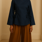 Jeune femme noire de dos portant une blouse bleu marine à manches longues évasées avec laçage côté et dos arrondi, associée à un pantalon large taille haute marron, dans un studio beige neutre.